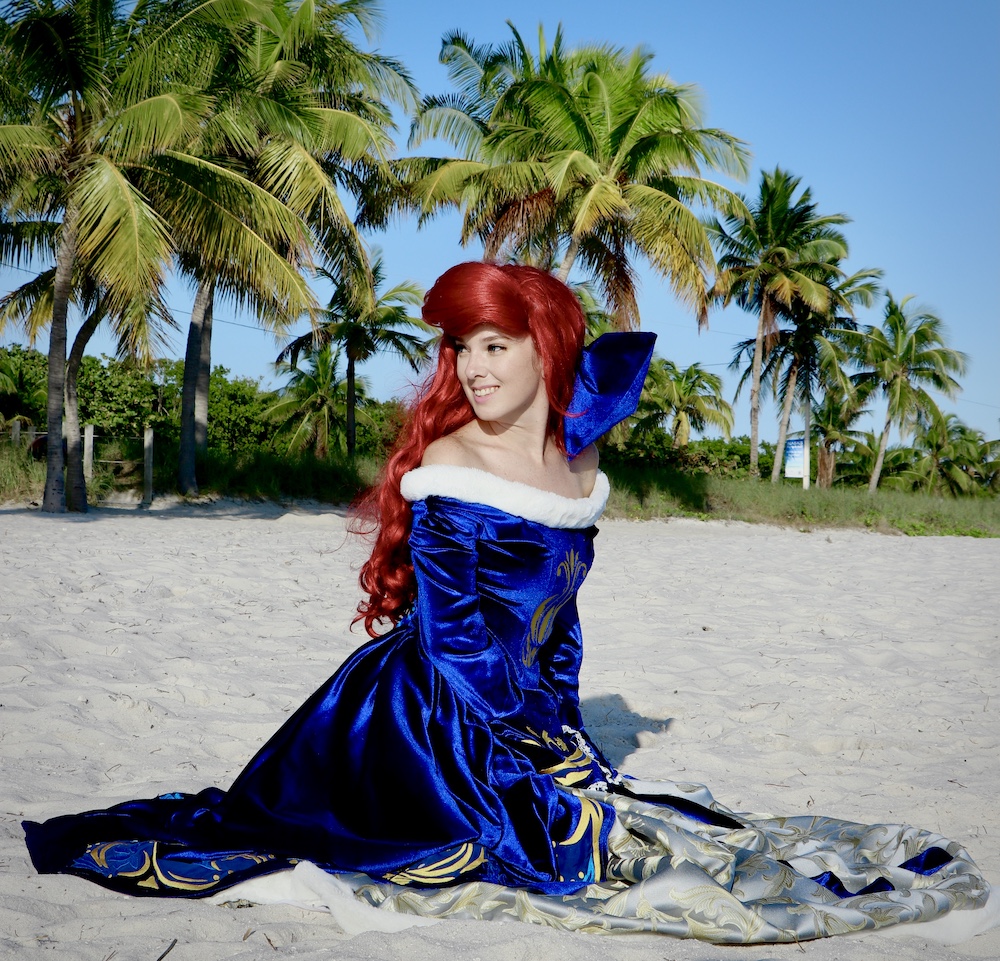 Christina dressed as Ariel (Christmas Edition 2020) on the Beach with a line of palm trees in the background.